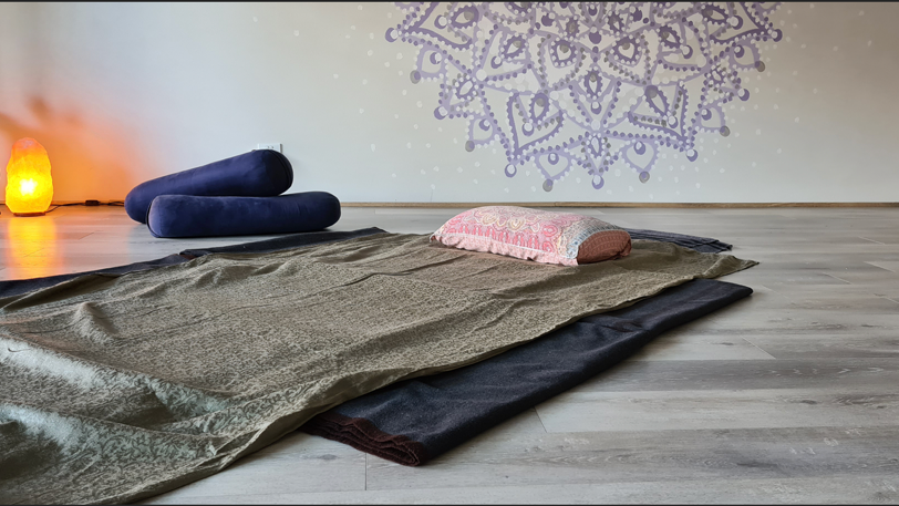 yoga blankets arranged on the floor with a pillow, bolsters in the background & a mandala on the wall